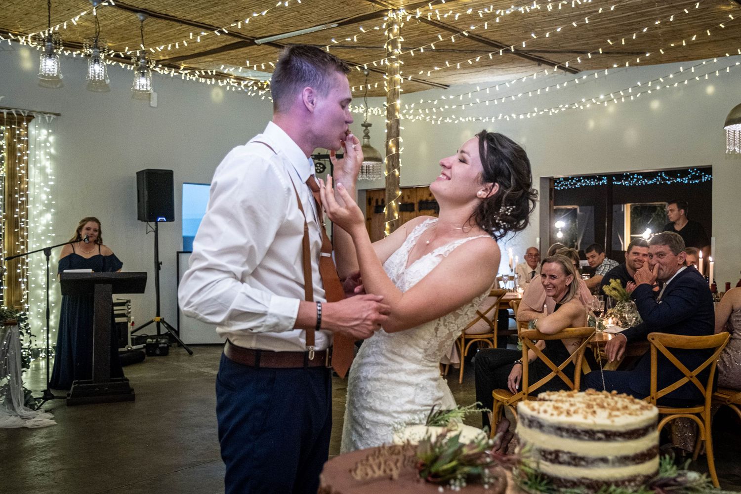 Bride playfully feeds groom cake frosting while guests watch and laugh at the wedding reception.