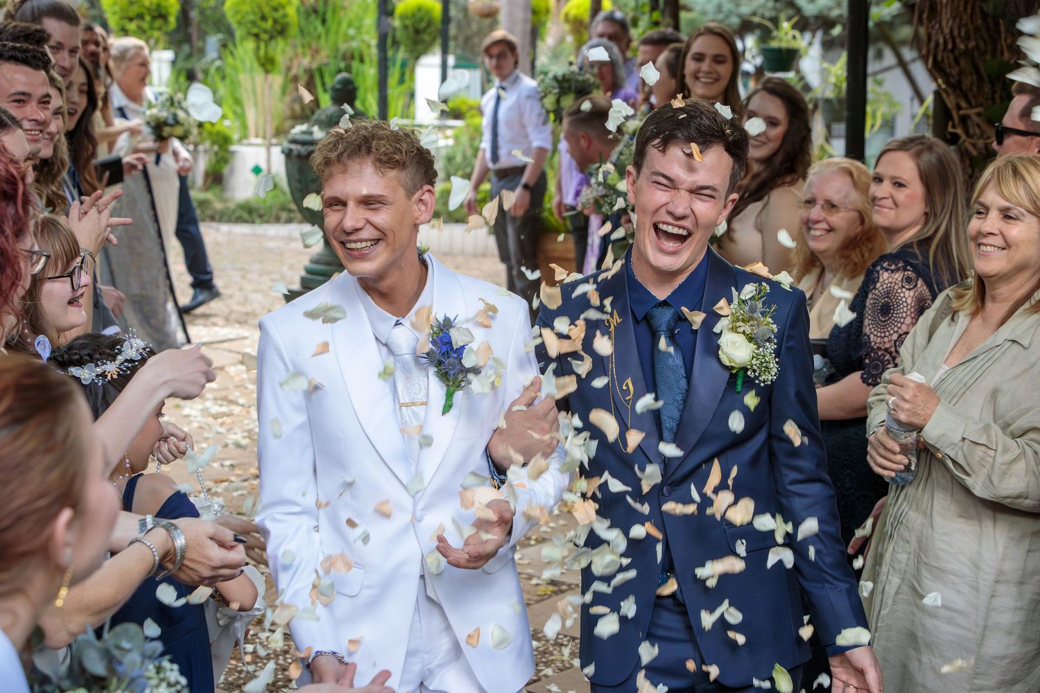 Two grooms laughing joyfully as guests throw flower petal confetti at outdoor wedding ceremony.