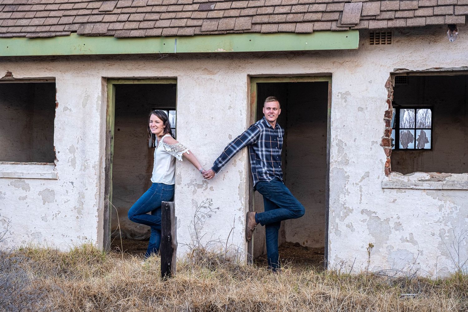 Couple posing playfully in doorways of abandoned rustic building, holding hands and balancing on one foot.