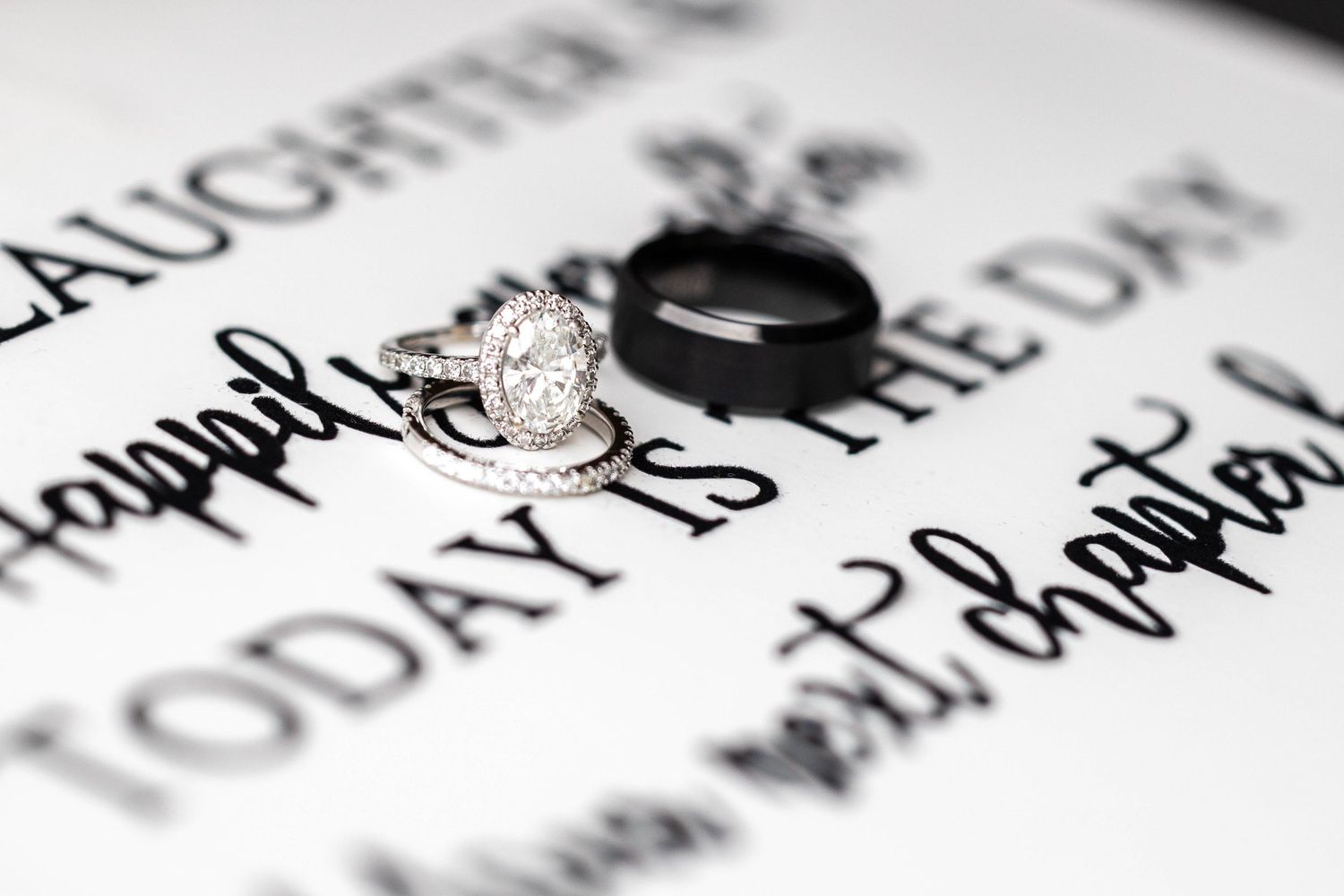 Diamond oval halo engagement ring and black wedding band on decorative calligraphy sign.