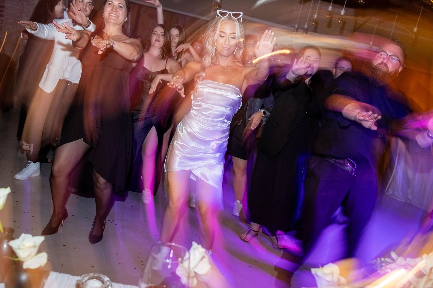 Bride in white dress dancing energetically with wedding guests under purple lights at reception.