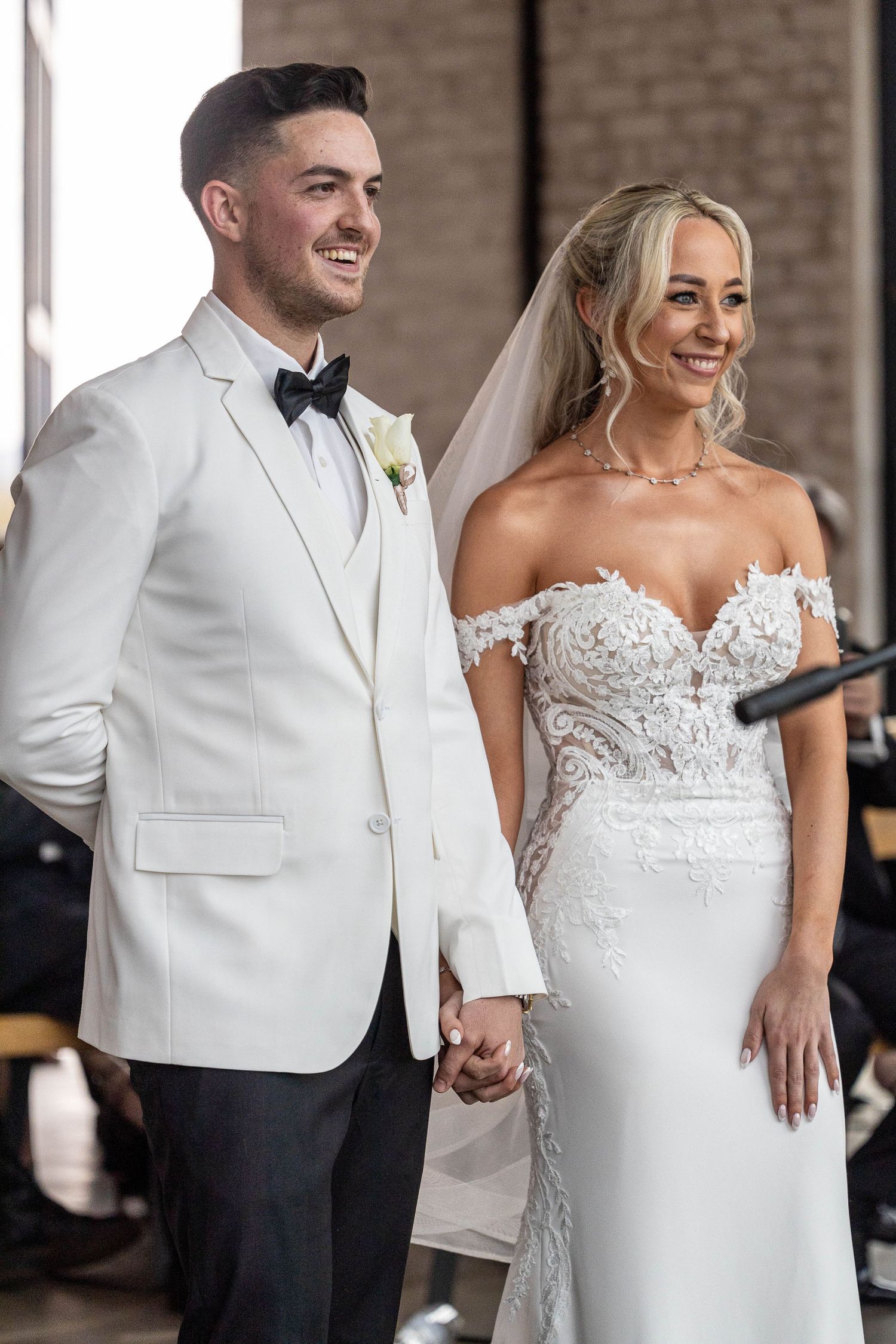 A joyful bride and groom in white wedding attire smile and hold hands at the wedding ceremony altar.