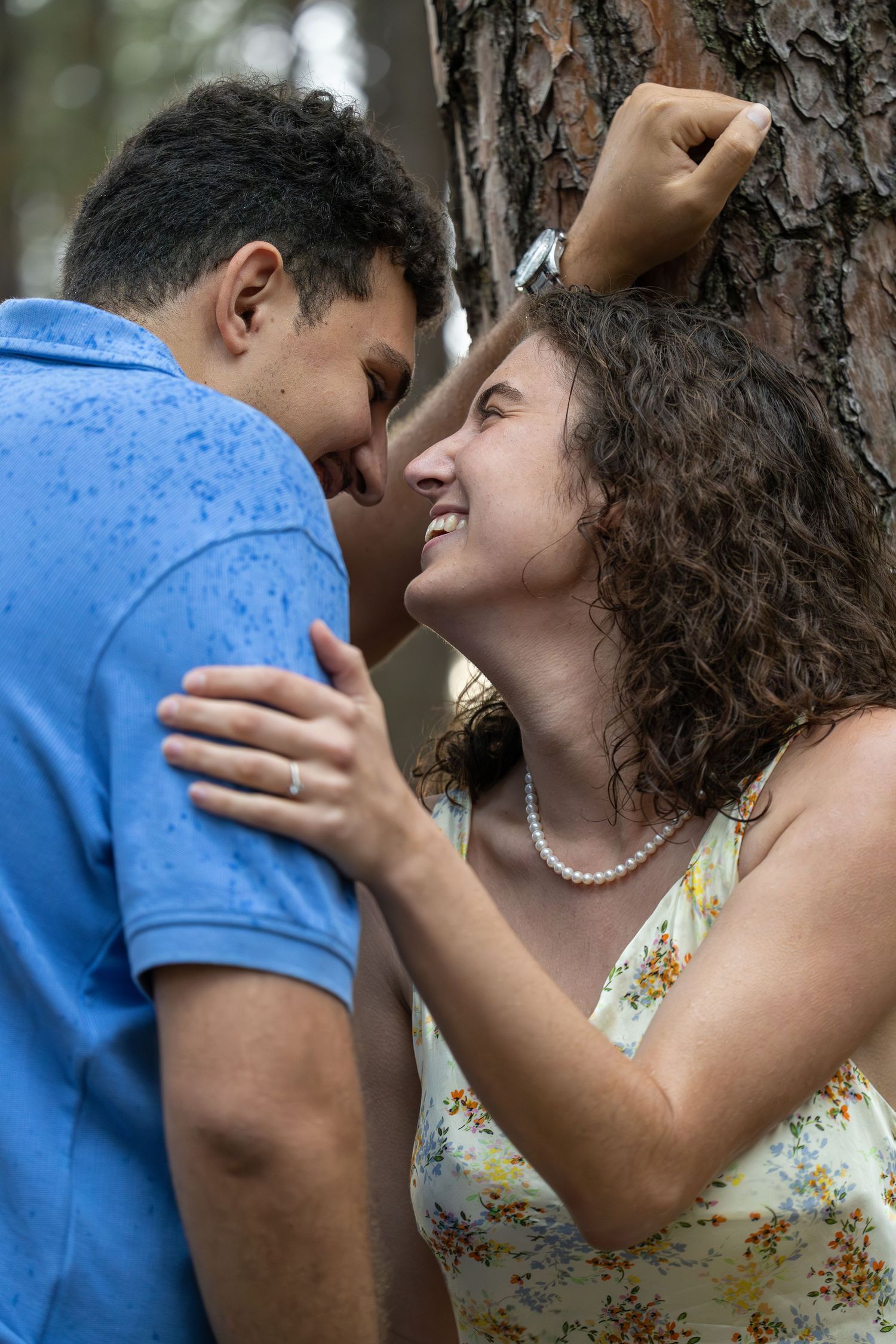 A young couple shares a romantic moment leaning against a tree, the woman smiling joyfully in a floral dress.