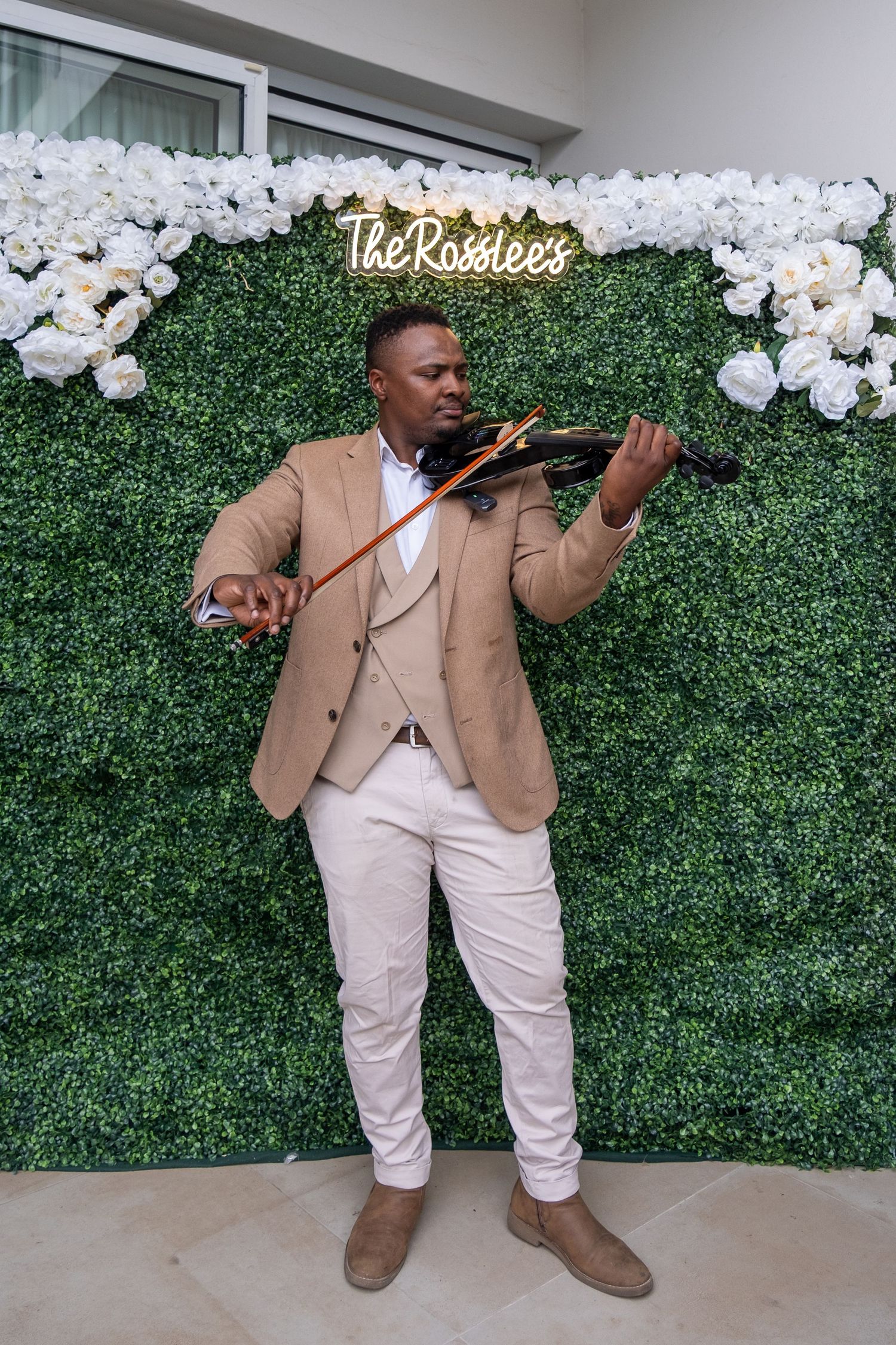 Musician in tan blazer plays electric violin in front of floral wall at The Rosslees wedding.