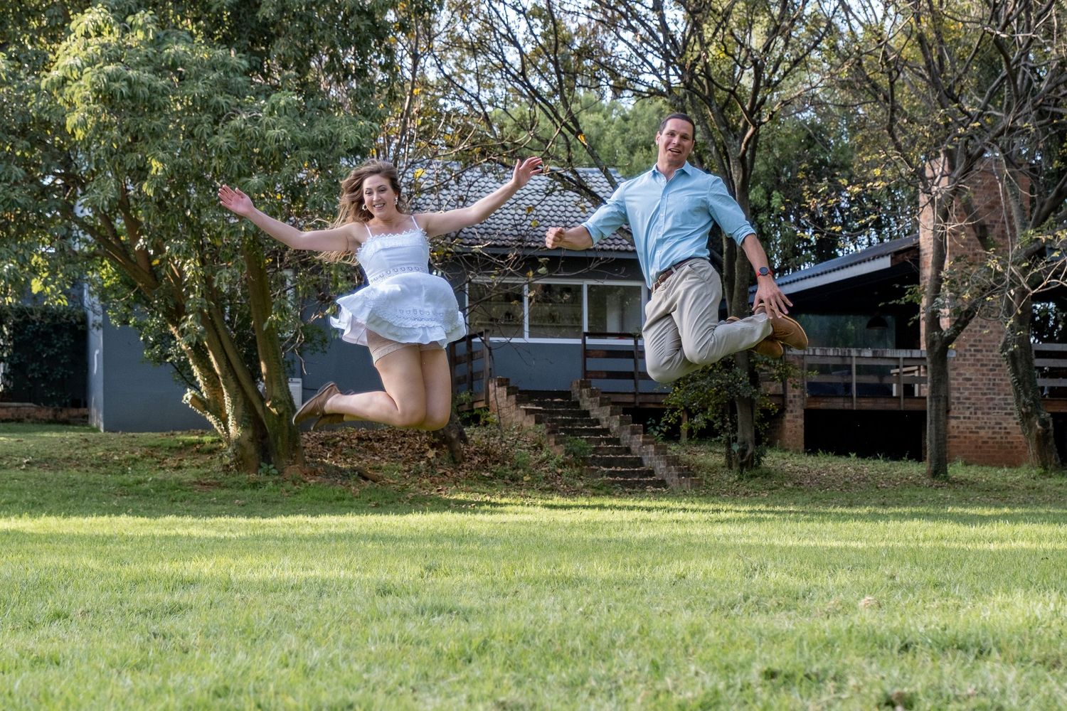 A happy couple jumps together in front of a charming house surrounded by trees.