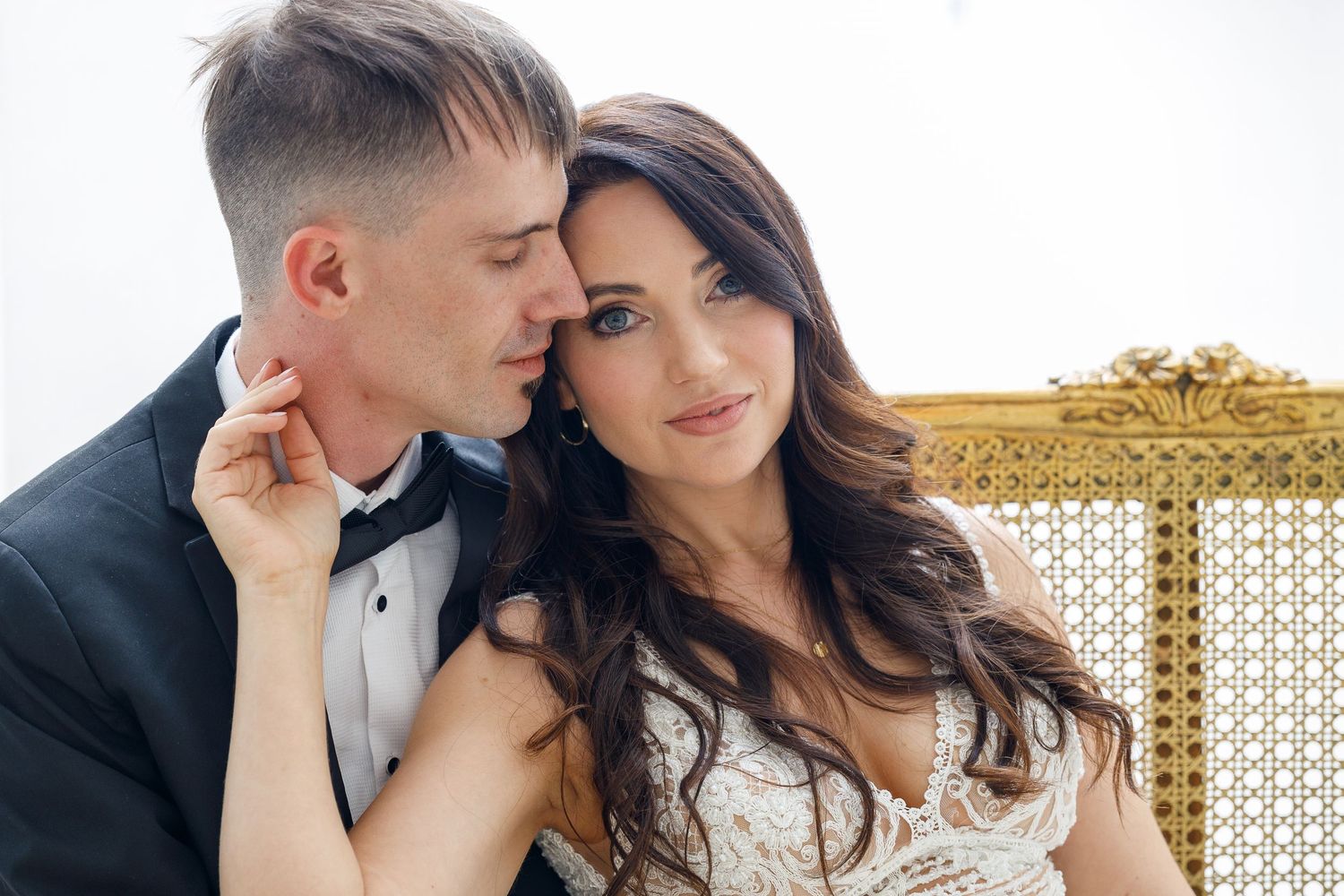 A bride in a lace wedding dress and groom in a black tuxedo share an intimate moment on an ornate gold chair.