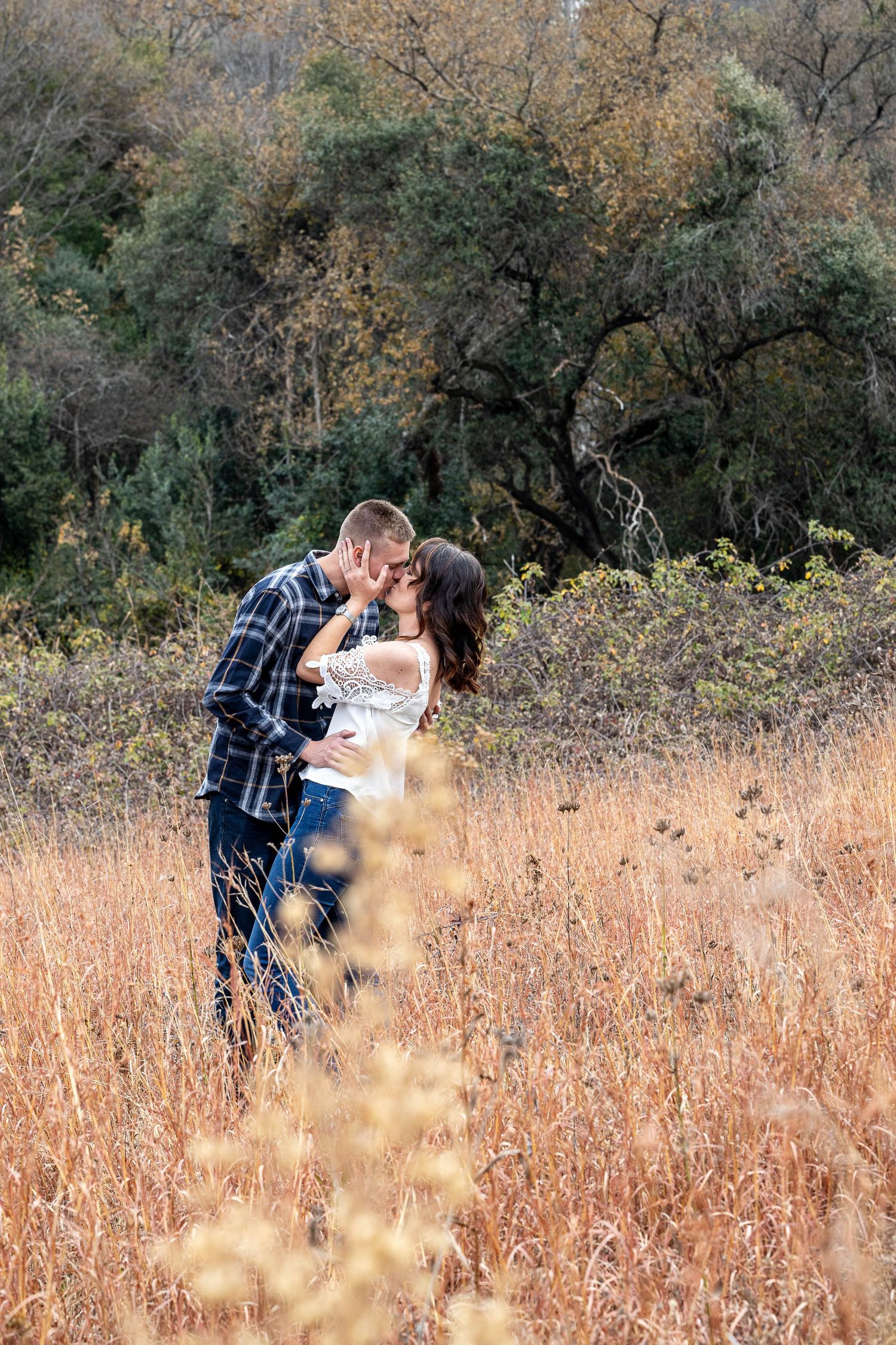 Couple shares passionate kiss in golden autumn field with lush trees in the background.