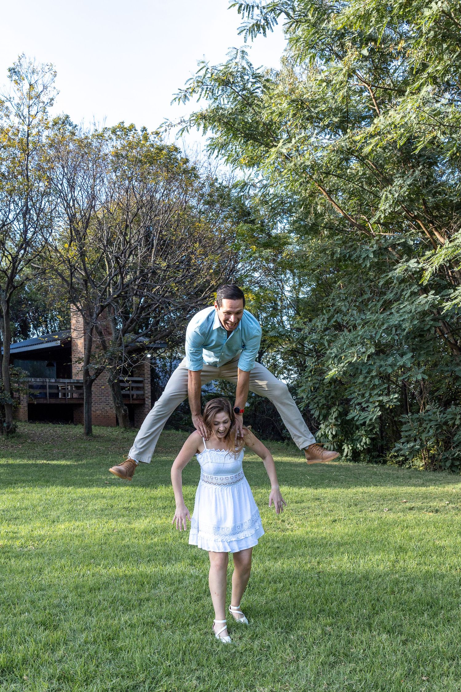 Man leaping playfully over a woman in a white dress on a green lawn during an engagement shoot.