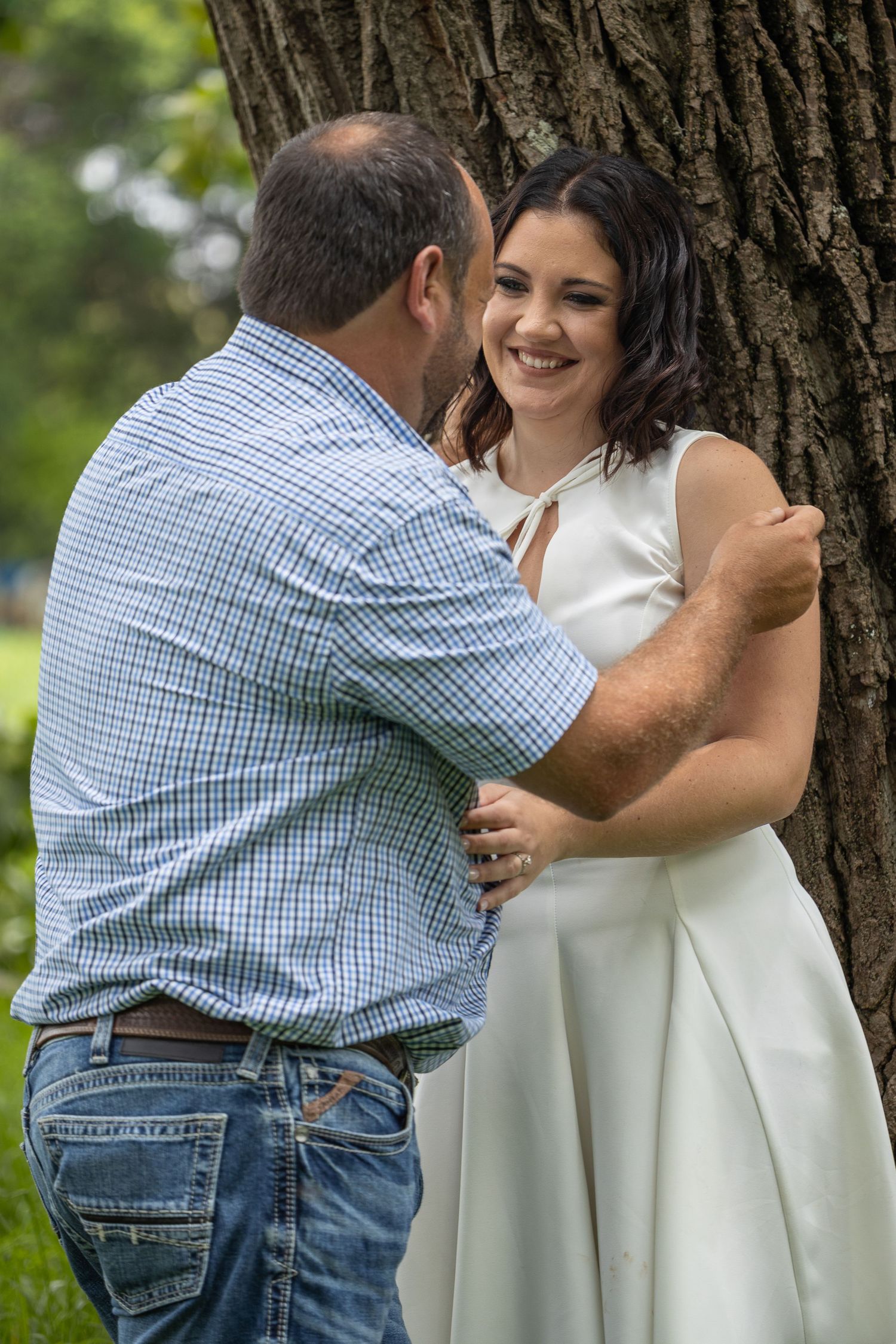 A couple shares a tender embrace near a large tree in a green park, the woman smiling brightly.