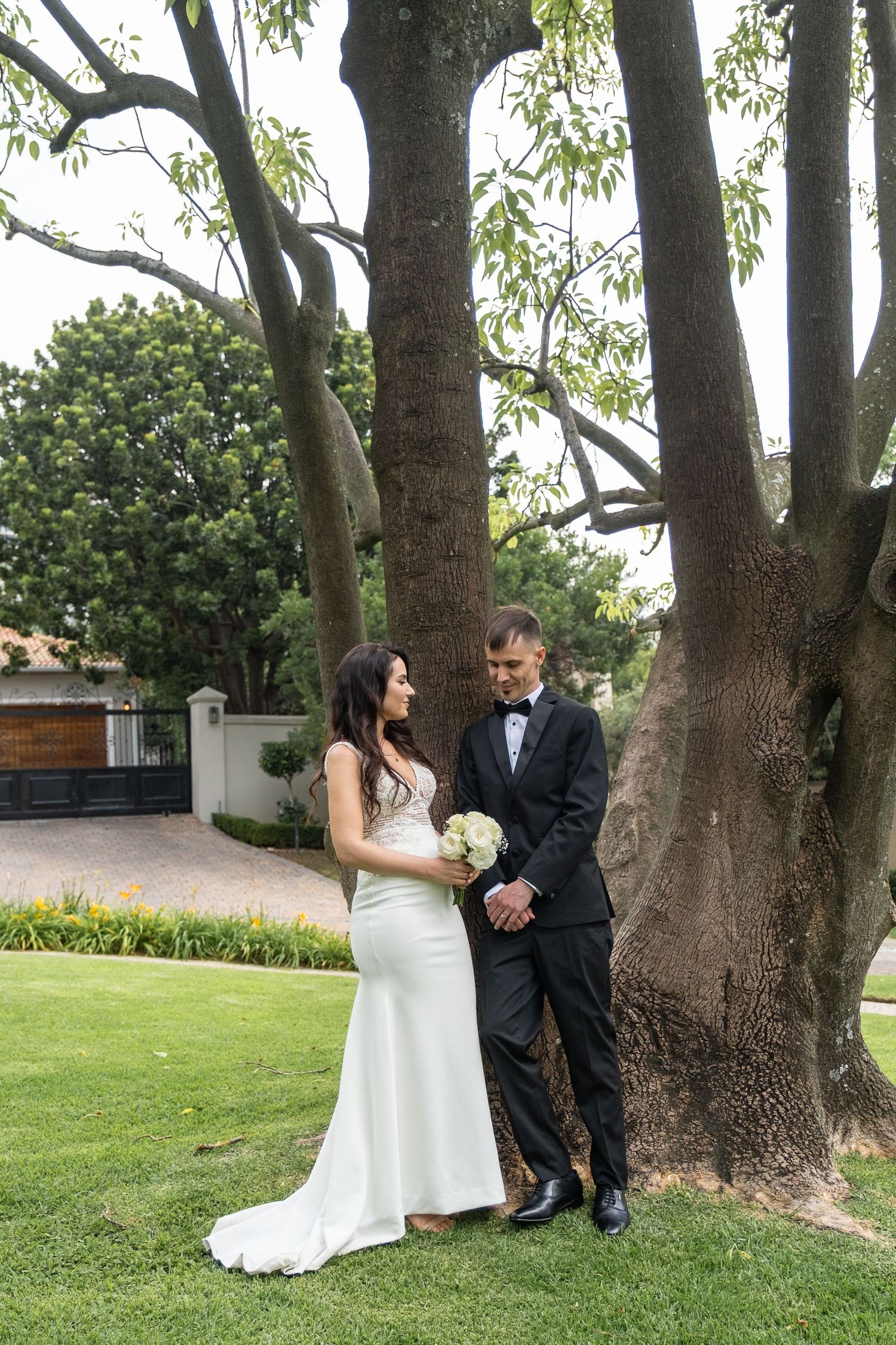 A bride in a white fitted gown and groom in a black tuxedo stand together under a large tree on a green lawn.