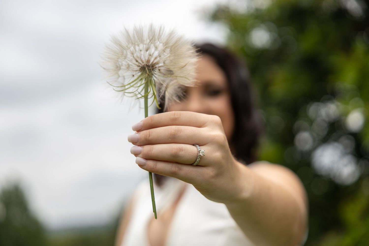 Woman in white dress holding a large dandelion flower with an engagement ring visible.