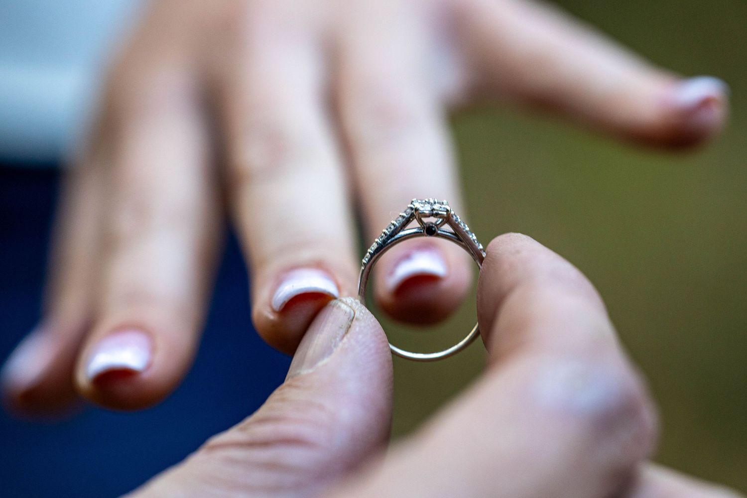 Close-up of a diamond engagement ring being held between two fingers outdoors.