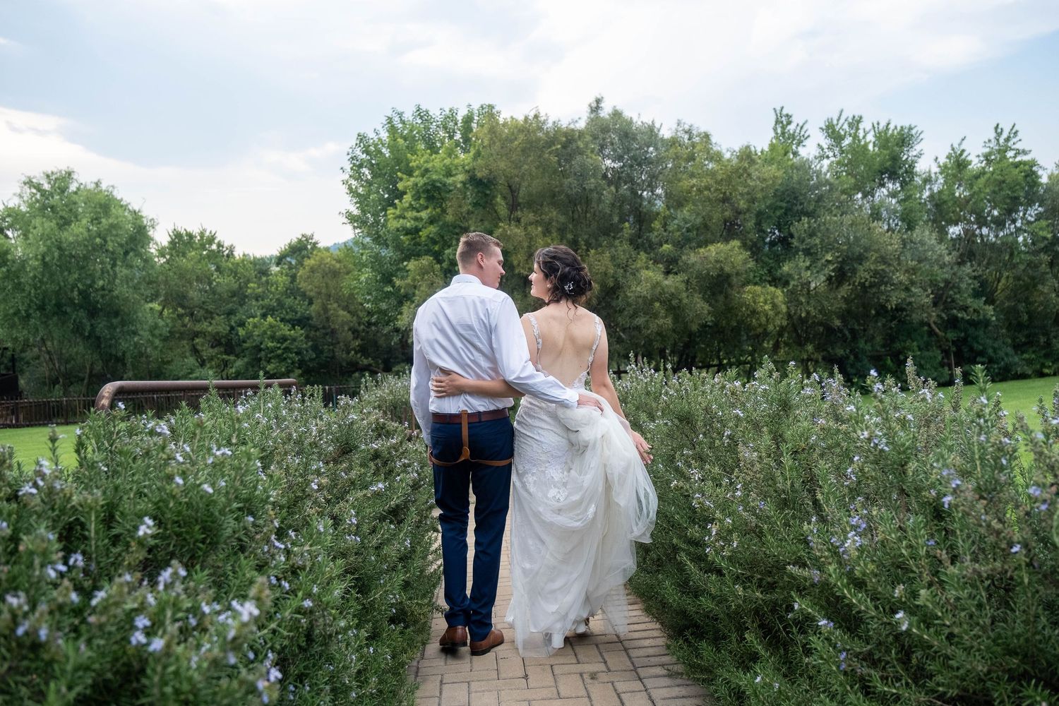 Bride and groom walk hand-in-hand along a garden path surrounded by lush green shrubs and trees on their wedding day.