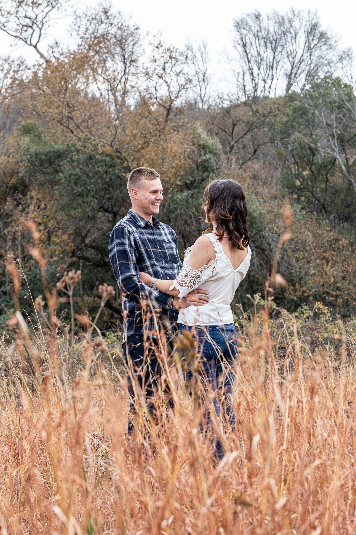 Smiling couple stands together in golden grass field surrounded by colorful autumn foliage trees.