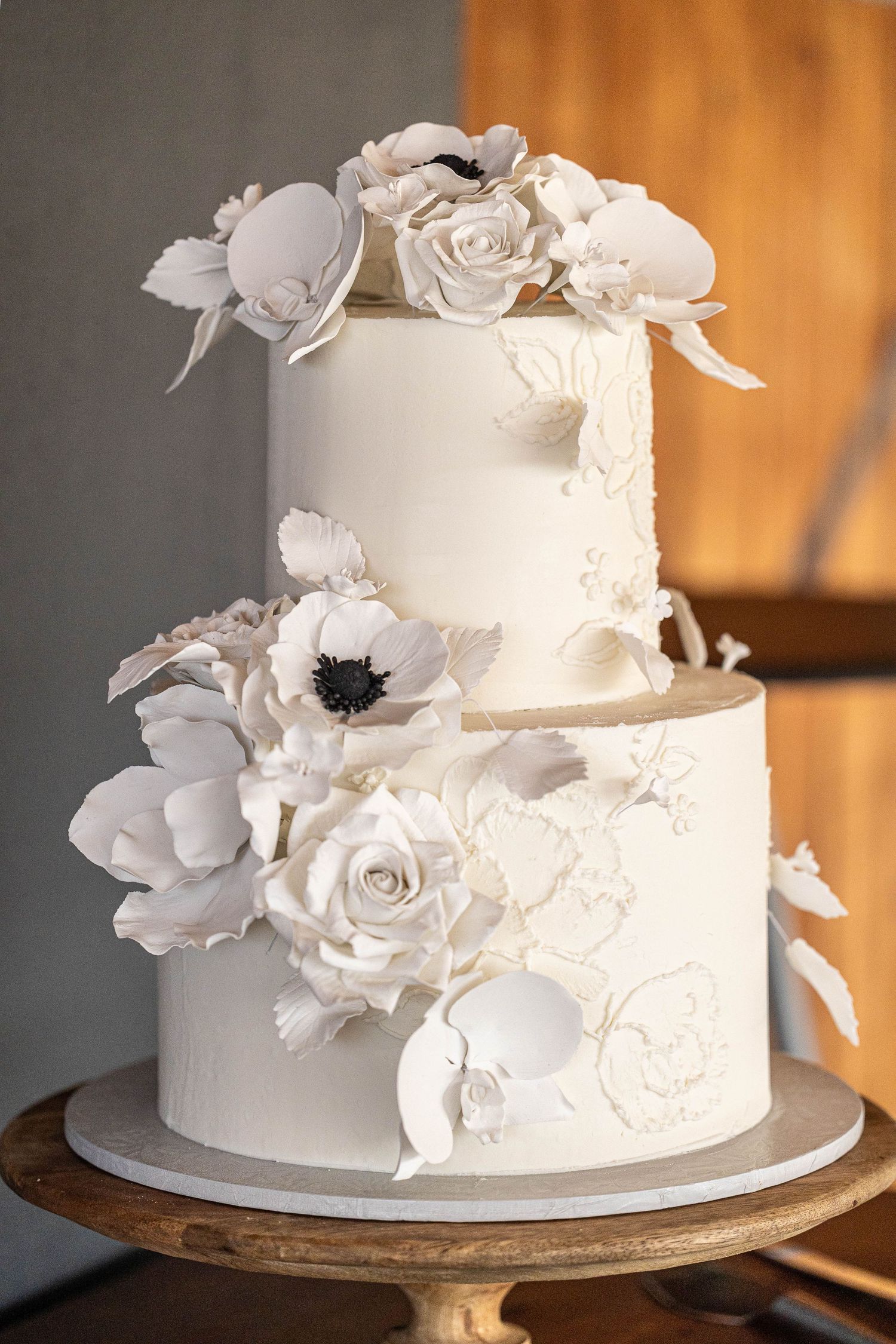 Elegant two-tier white wedding cake decorated with sugar flowers including roses and anemones on wooden stand.