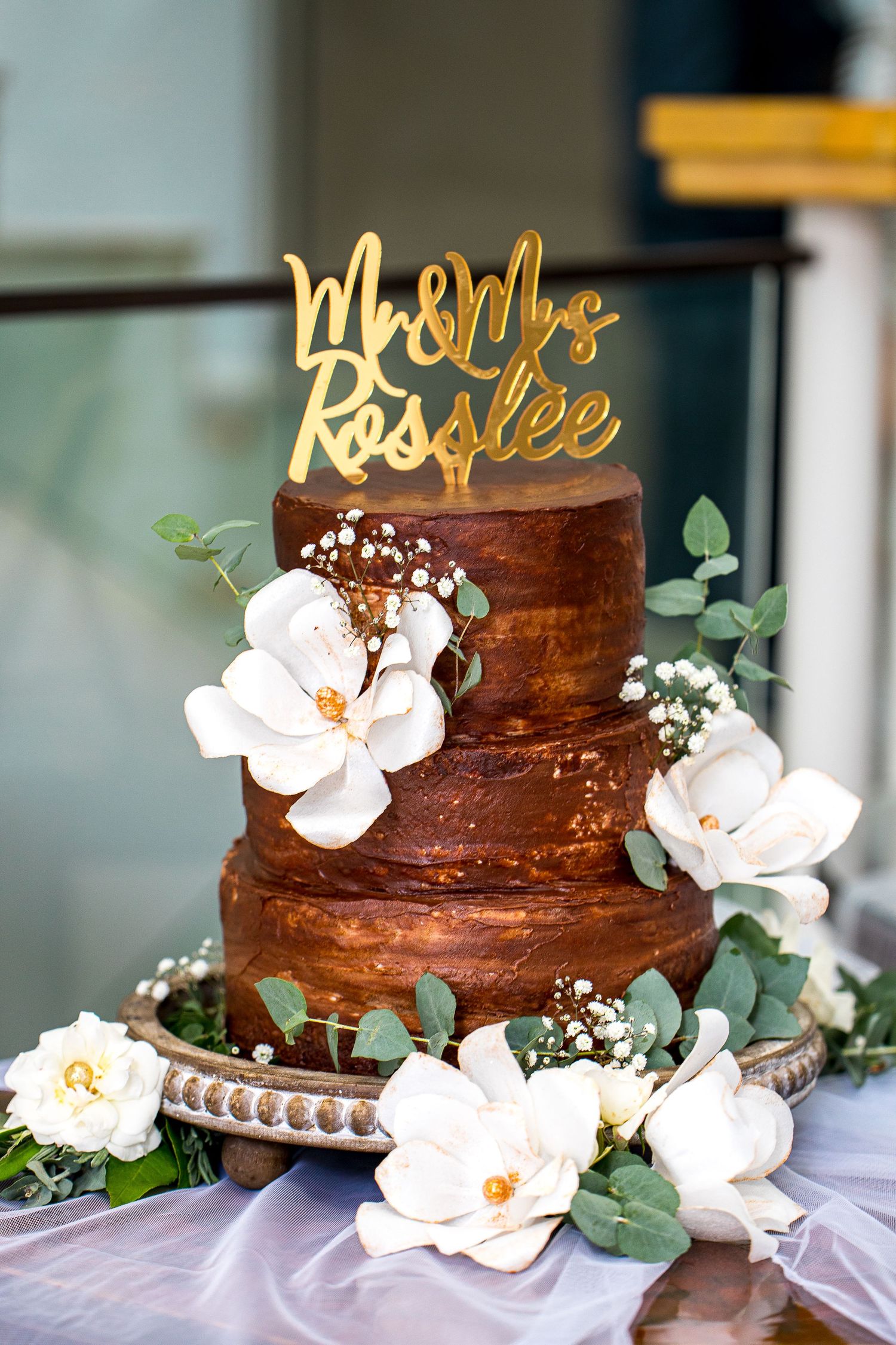 Three-tier chocolate wedding cake with white magnolias and Mr & Mrs Rosslee gold cake topper.