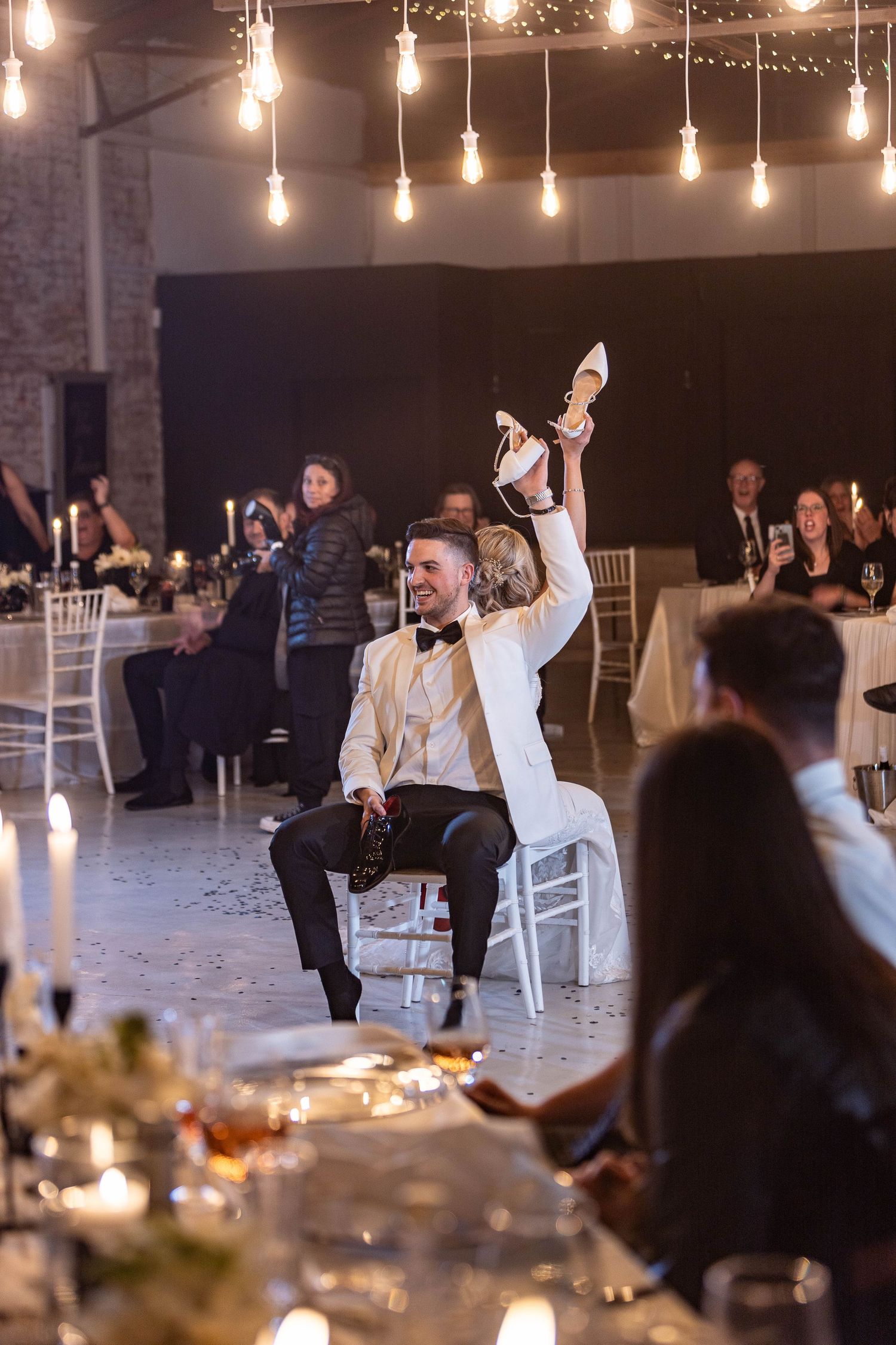 Bride raises shoe during fun wedding game while groom sits in chair at elegant candlelit reception venue.