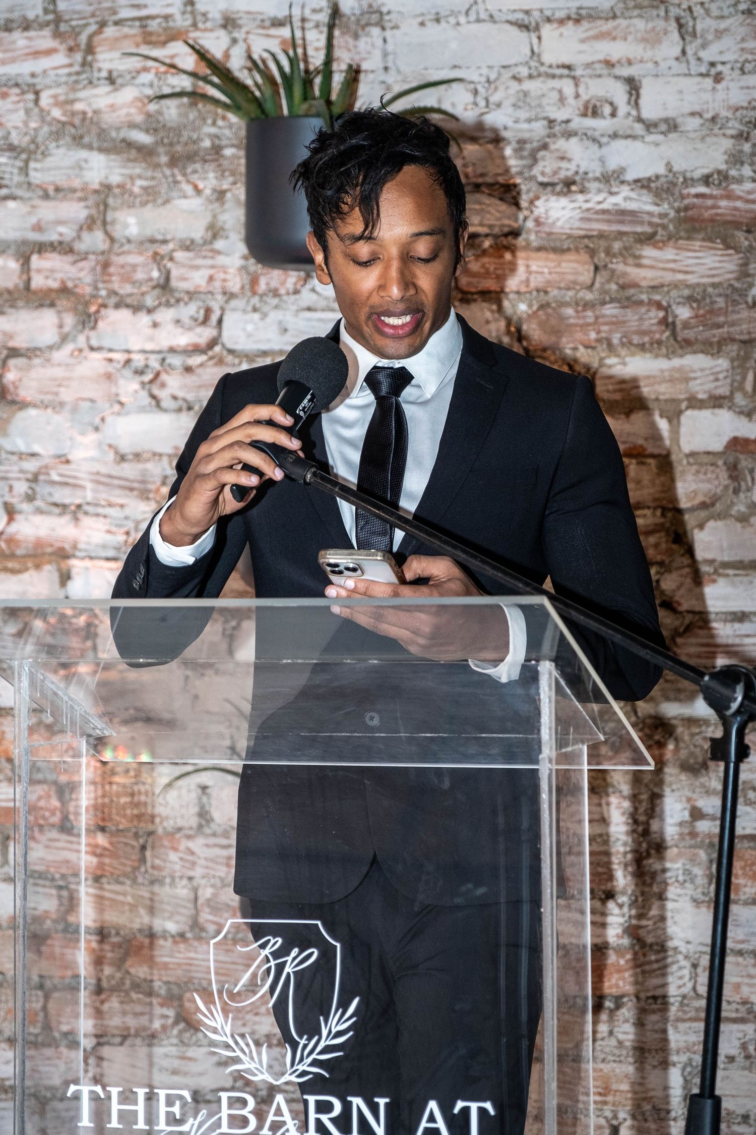 Elegantly dressed man delivers speech at clear acrylic podium at The Barn At wedding venue.