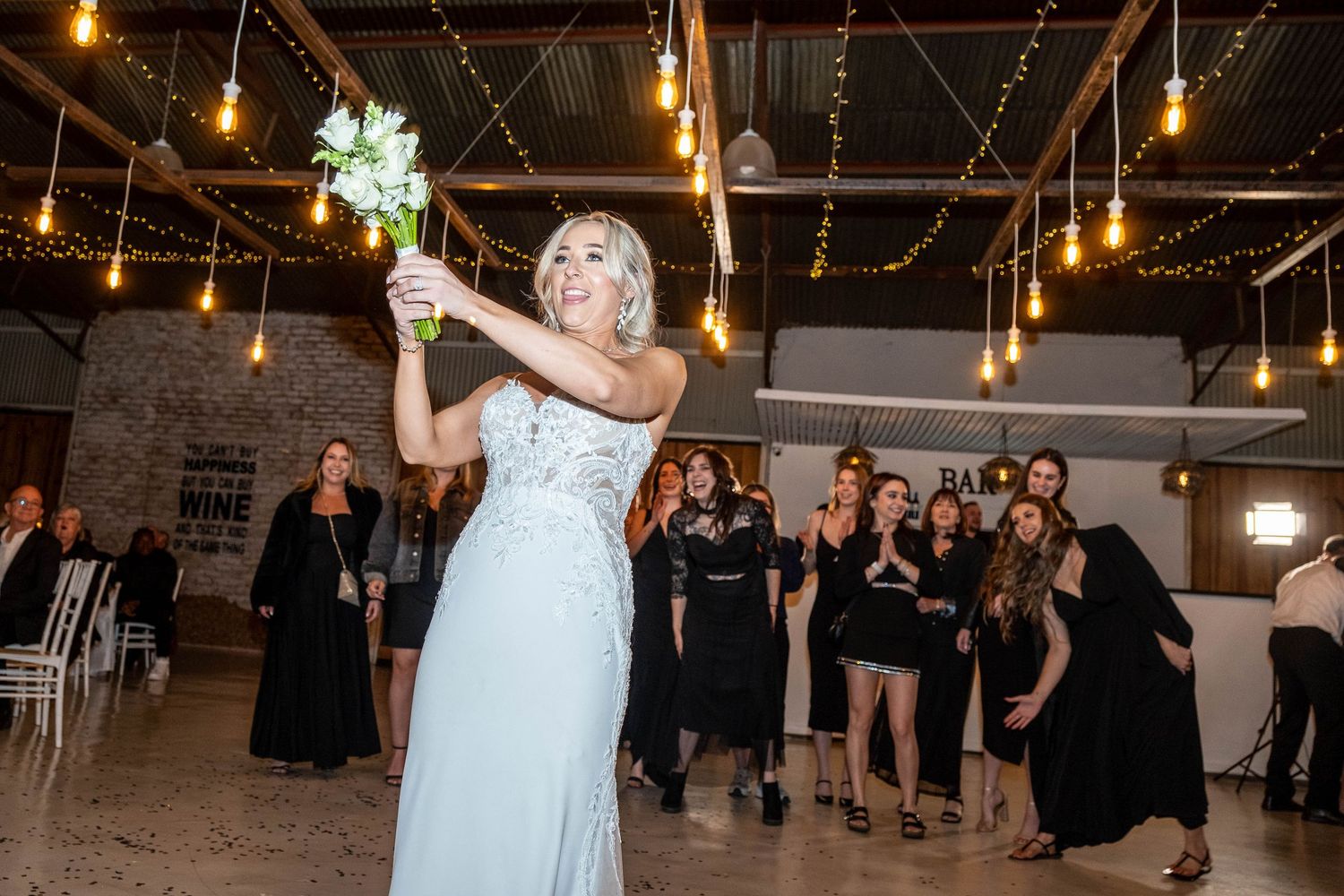 Bride in light blue gown tossing wedding bouquet to guests at indoor reception with Edison bulb lighting.