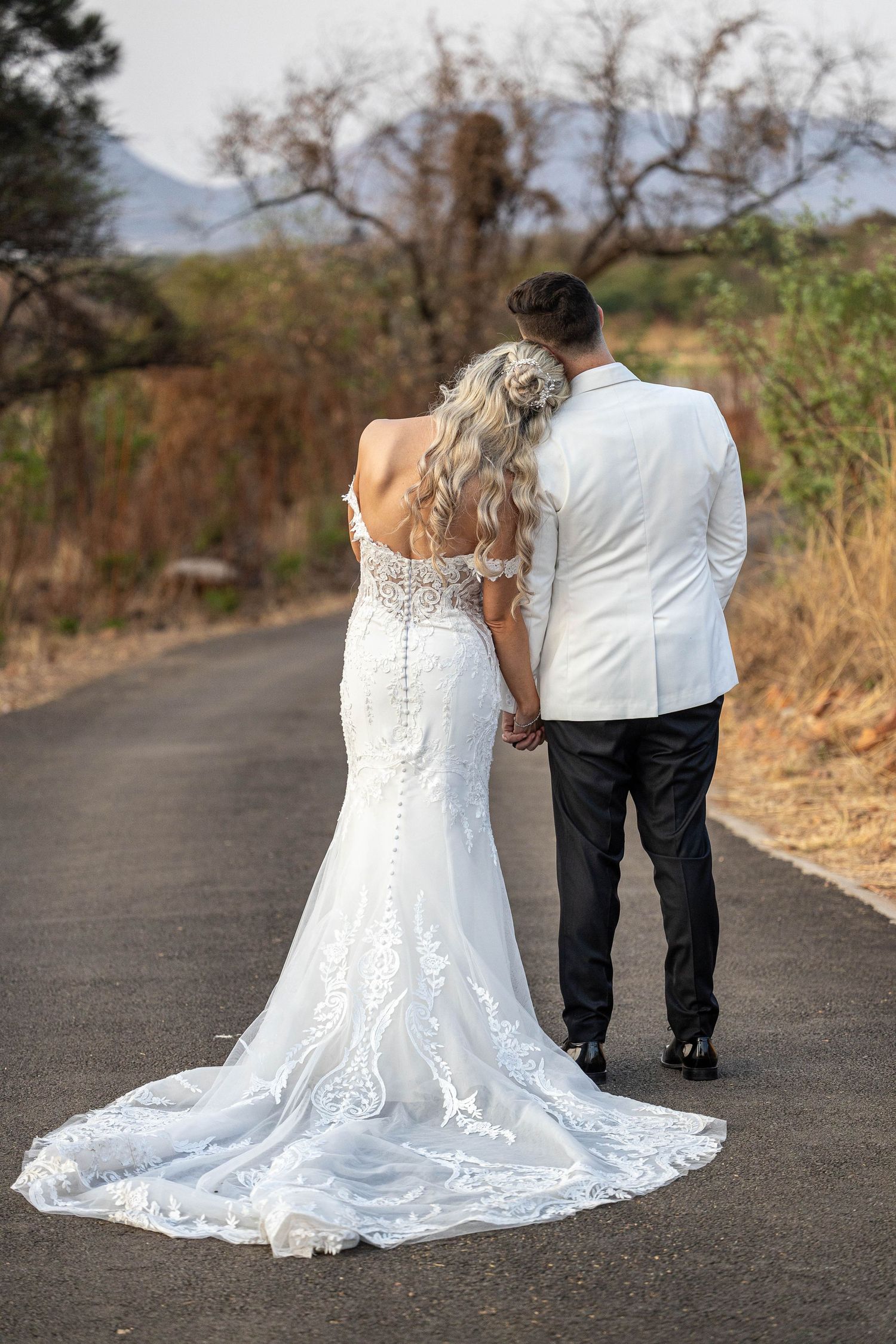 Bride and groom walking away down a country road, showcasing the bride's elegant lace gown and train.