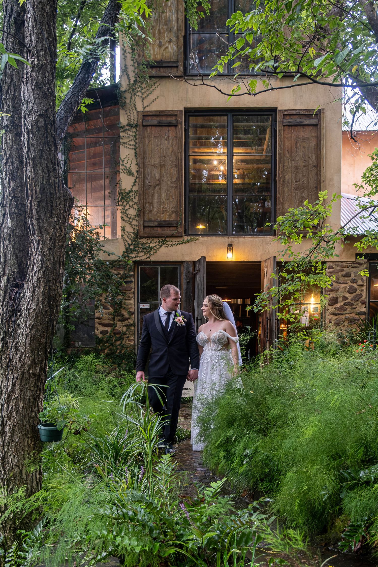 Bride and groom walking through a lush green garden courtyard outside a charming rustic stone building at twilight.