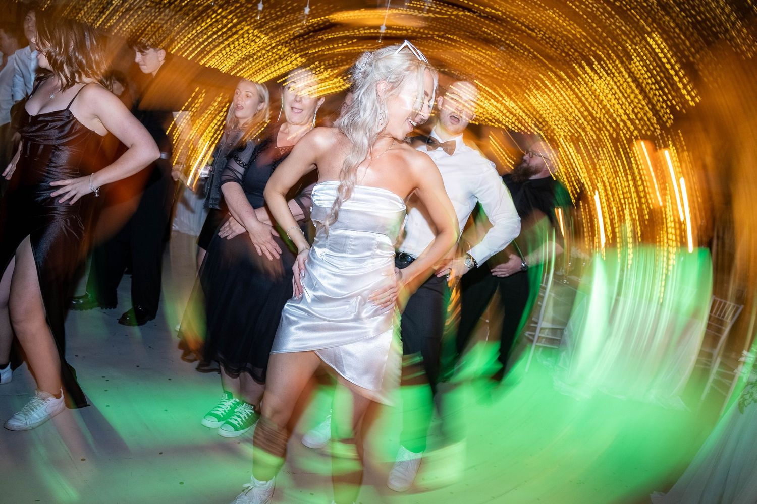 Bride in short white dress dancing energetically with guests under golden lights at wedding reception.