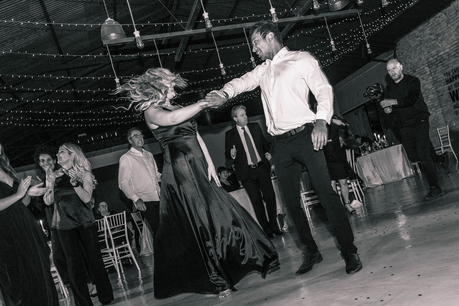 Couple shares energetic dance at wedding reception in black and white photo with guests watching.