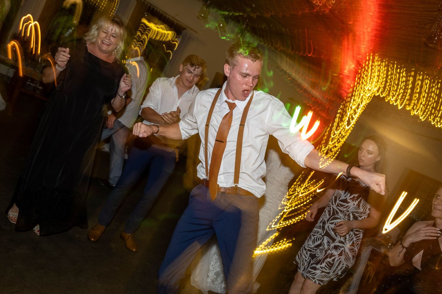 Groomsman dancing energetically with guests under swirling golden lights at wedding reception.