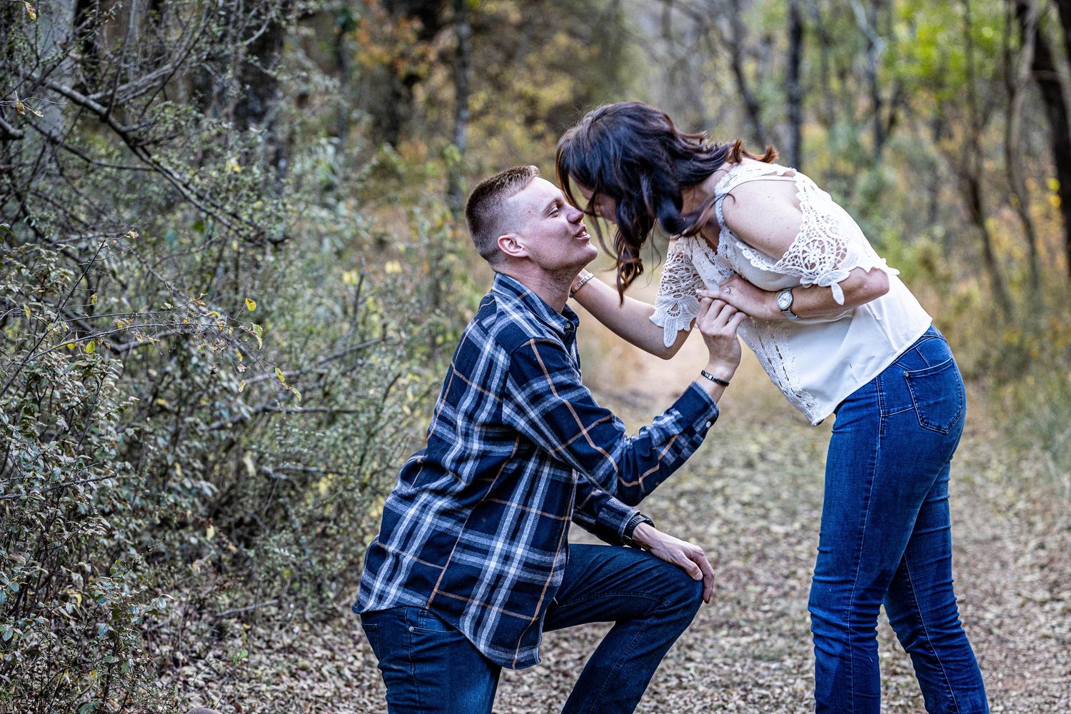 Couple sharing a kiss as man kneels on one knee during a romantic outdoor marriage proposal moment.