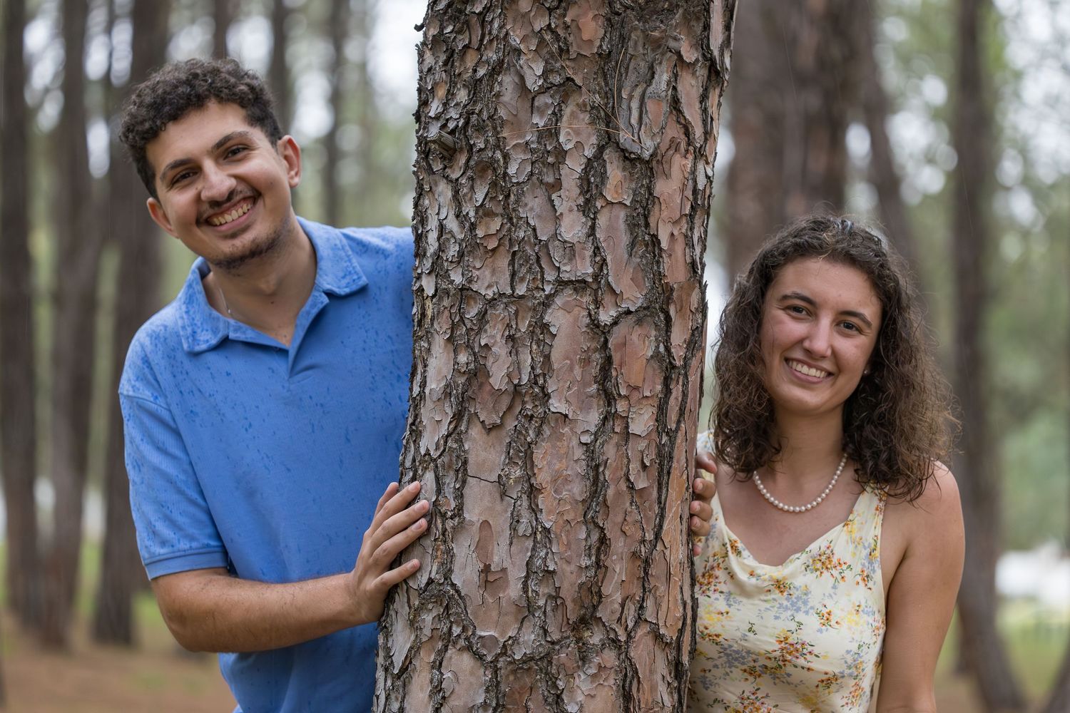 Smiling couple posing together on either side of a pine tree outdoors.