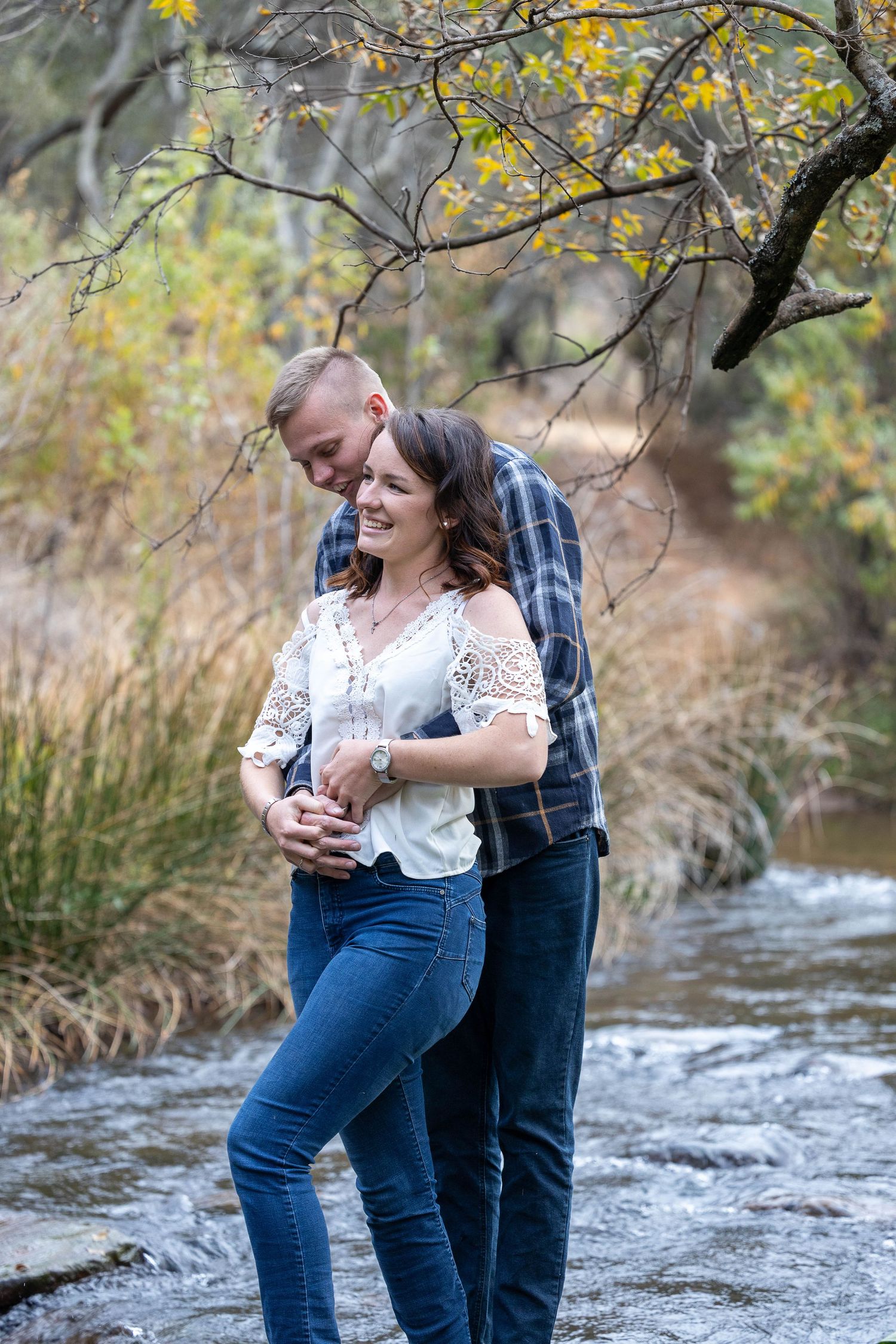 Happy couple embracing by a flowing creek surrounded by autumn foliage during outdoor engagement photos.