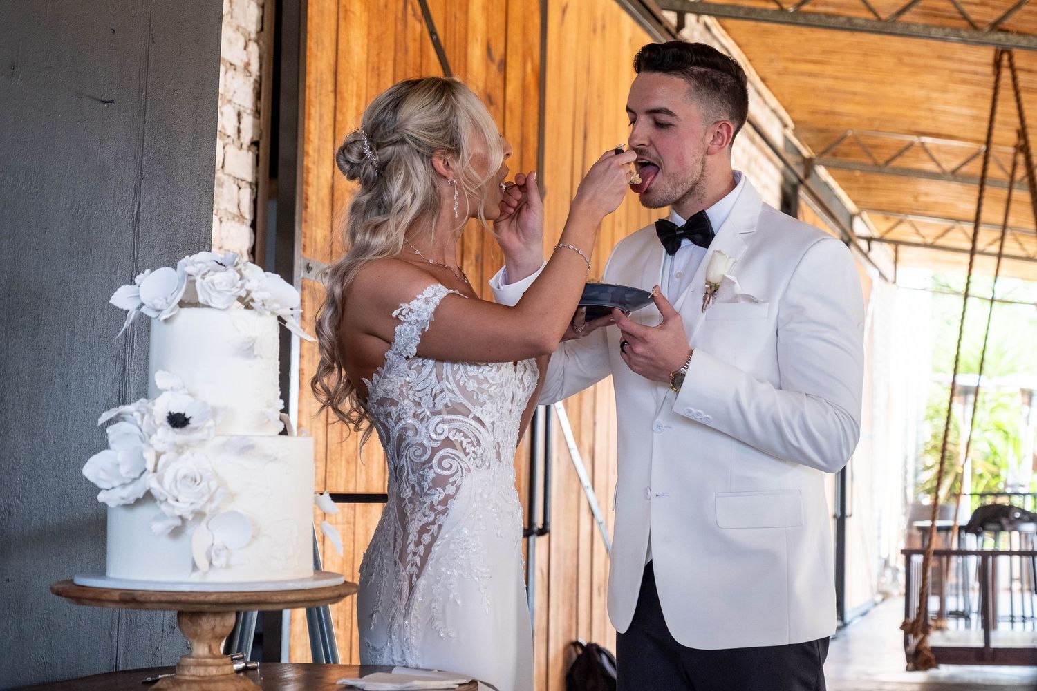 Bride feeding wedding cake to groom during reception near elegant white floral tiered cake display.