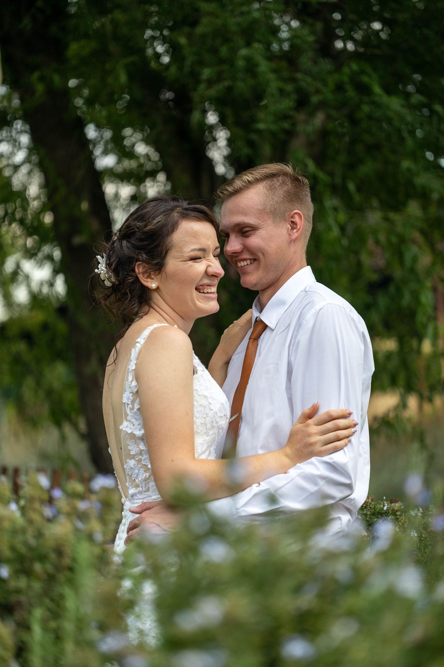 Laughing bride and smiling groom embrace joyfully outdoors surrounded by green garden foliage on their wedding day.