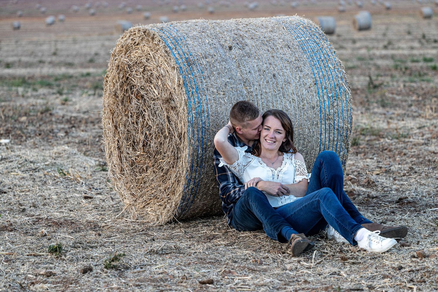 Couple sitting against a large hay bale in a harvested field, sharing a tender kiss on the cheek.