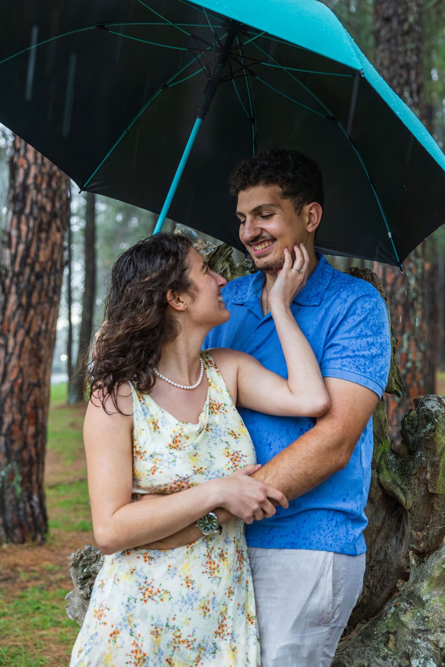 Happy couple sharing a romantic moment under a teal umbrella in a pine forest.