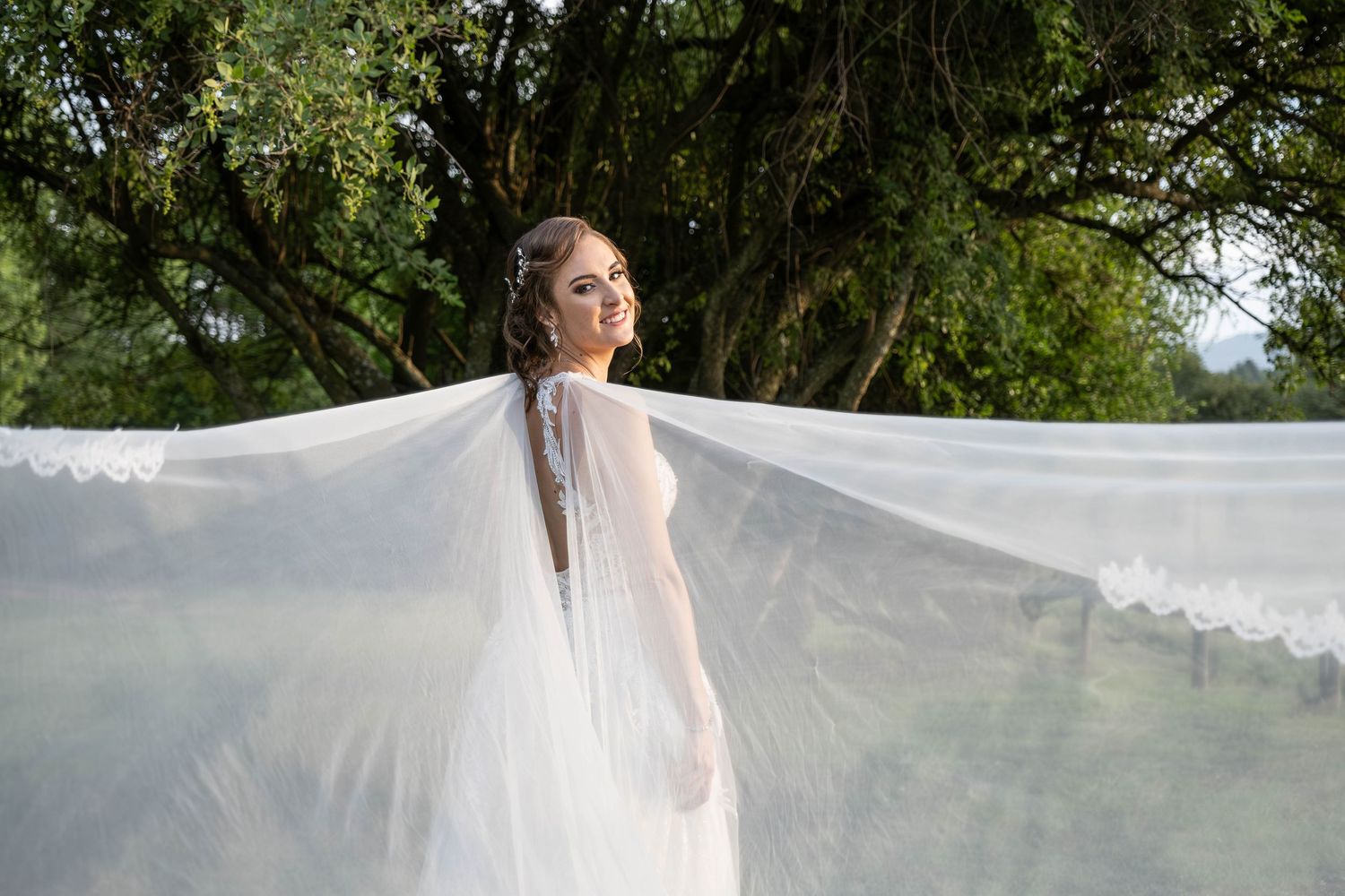 Bride in white gown with flowing lace-trimmed cathedral veil spread wide in outdoor garden setting.