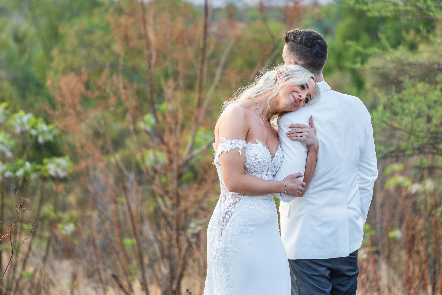 Bride in off-shoulder lace gown resting her head on groom's chest in a tender outdoor wedding moment.
