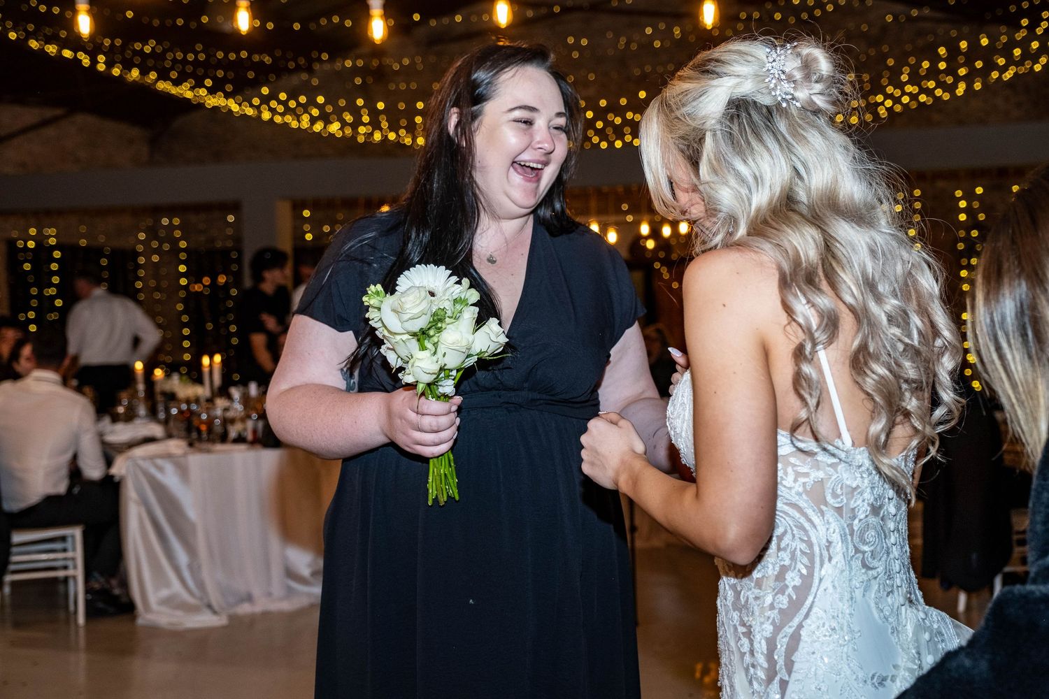 Wedding guest in black dress laughing while holding bouquet after catching it at reception.