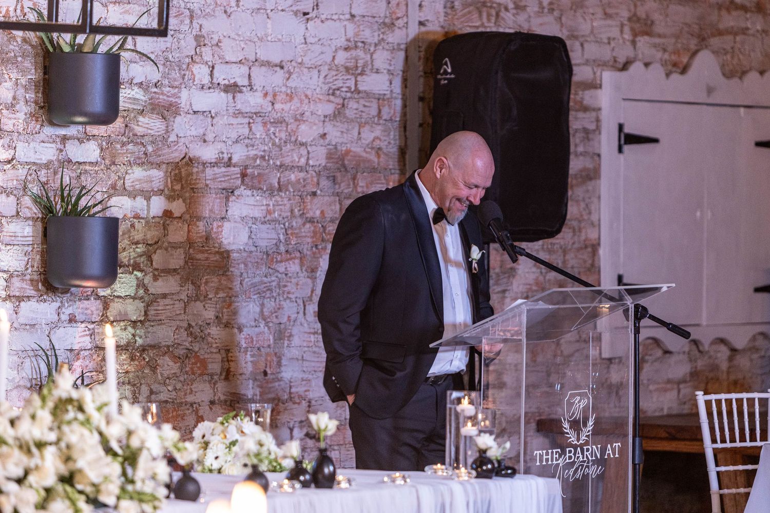 Bald man in tuxedo delivers heartfelt speech at clear podium during elegant wedding reception.