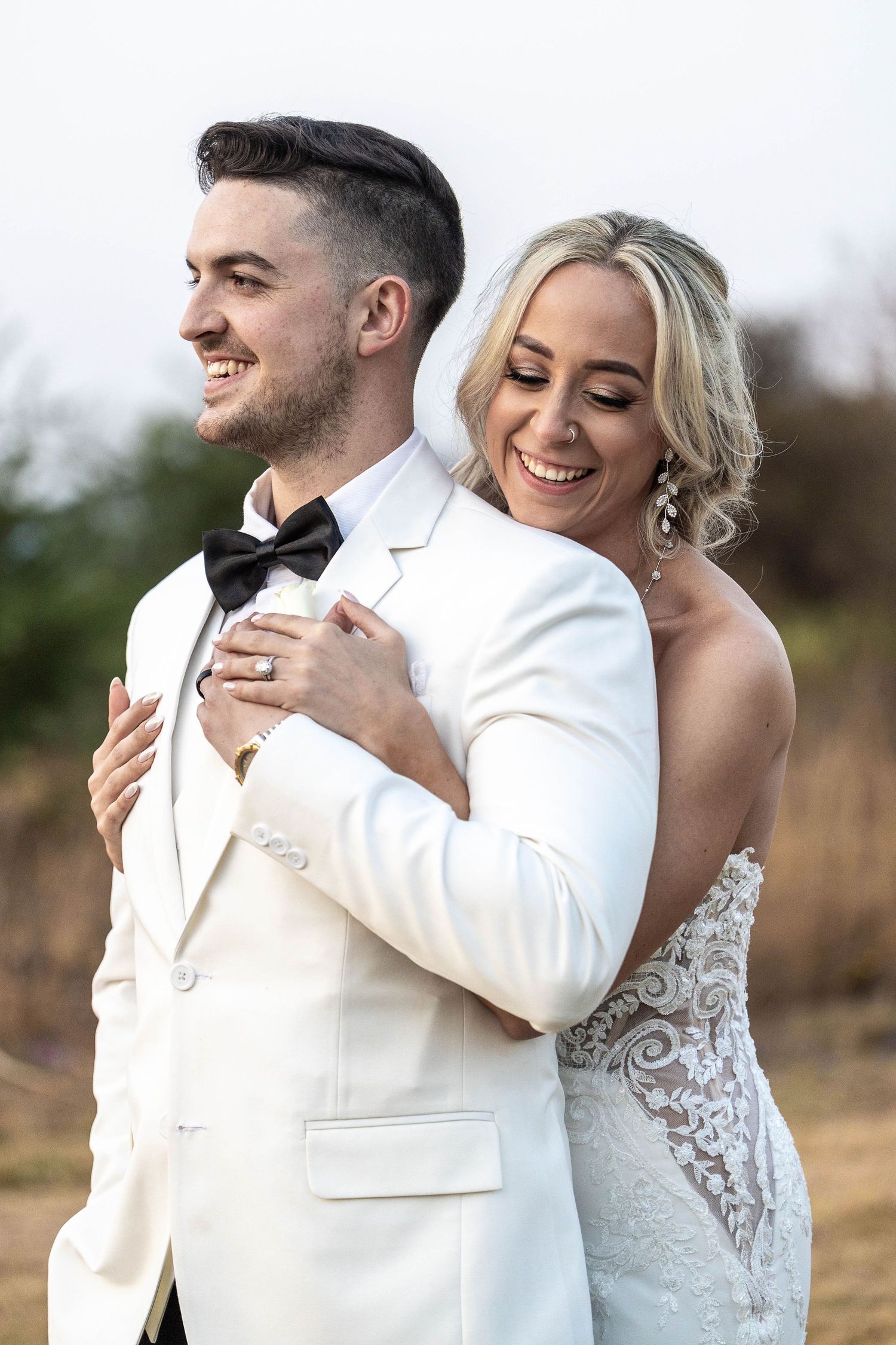 Laughing bride embracing groom from behind outdoors, both in elegant white wedding attire.