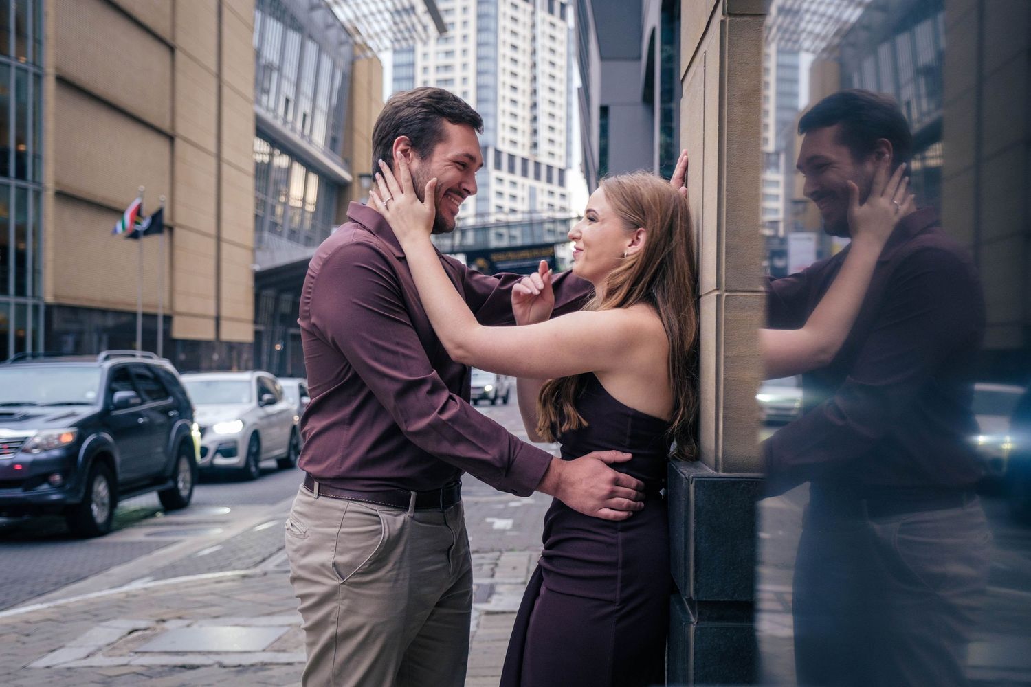 Couple shares romantic moment on urban city street, woman in black dress touching man's face near glass building.