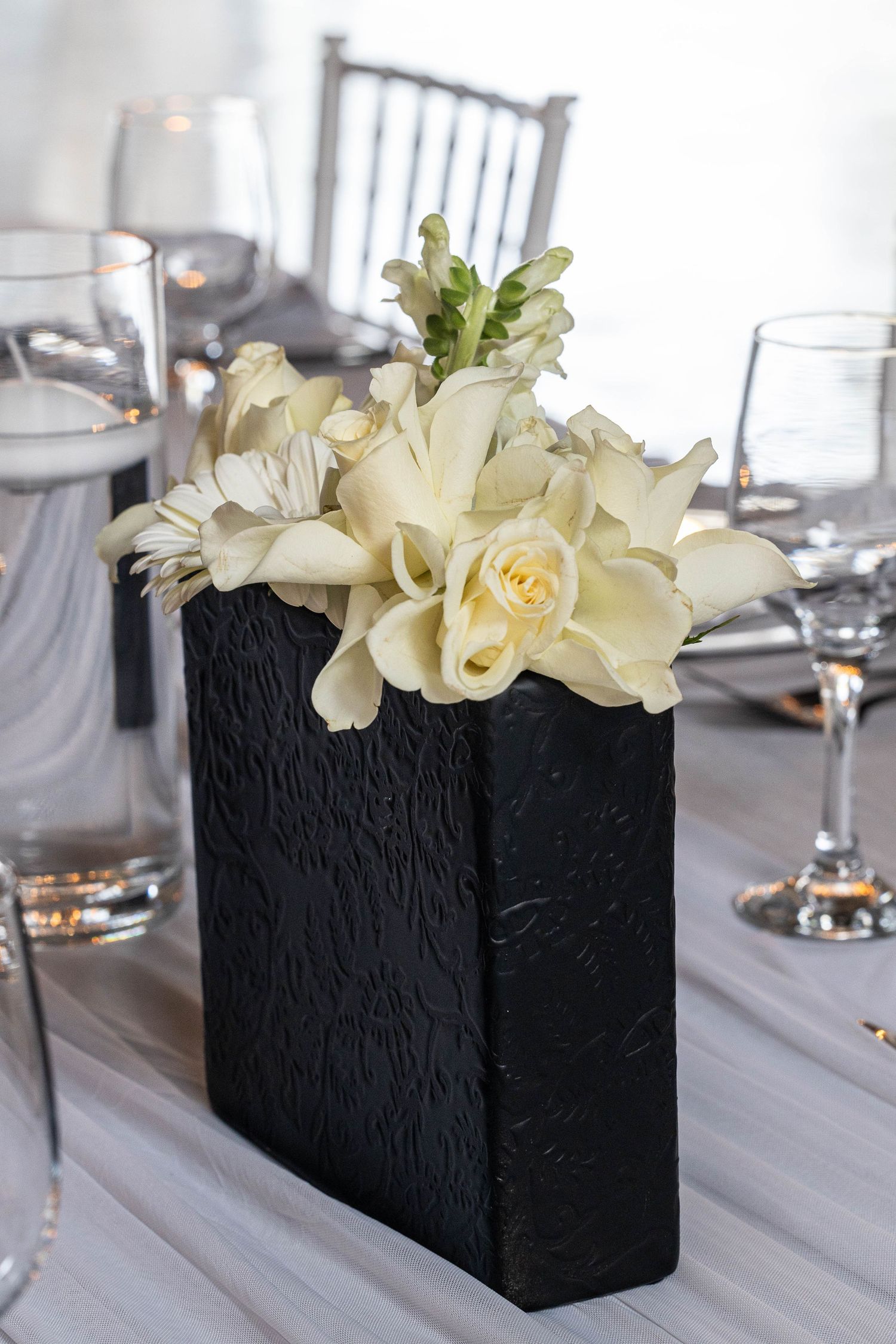 White roses and orchids arranged in black textured rectangular vase on elegant reception table.