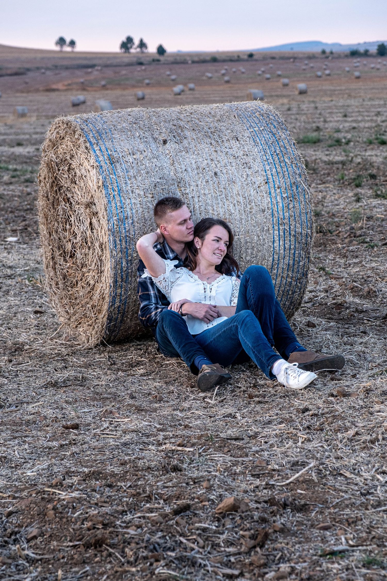 Happy couple sitting together against large round hay bale in harvested field at dusk, smiling warmly.