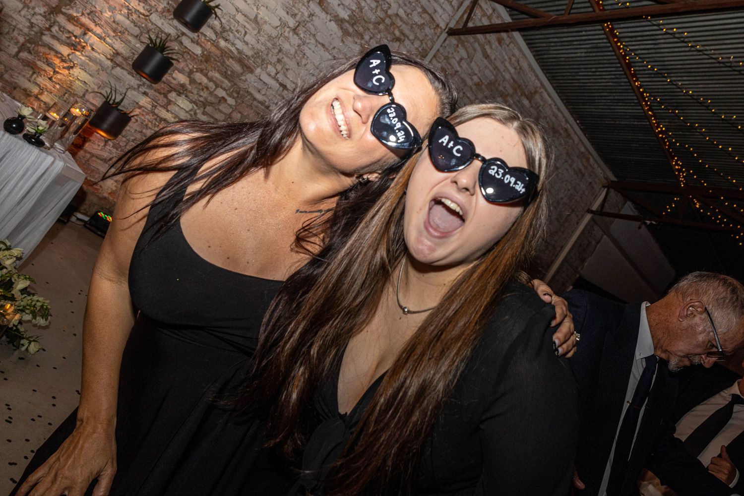 Two women wearing fun wedding sunglasses laughing together at a wedding reception party.