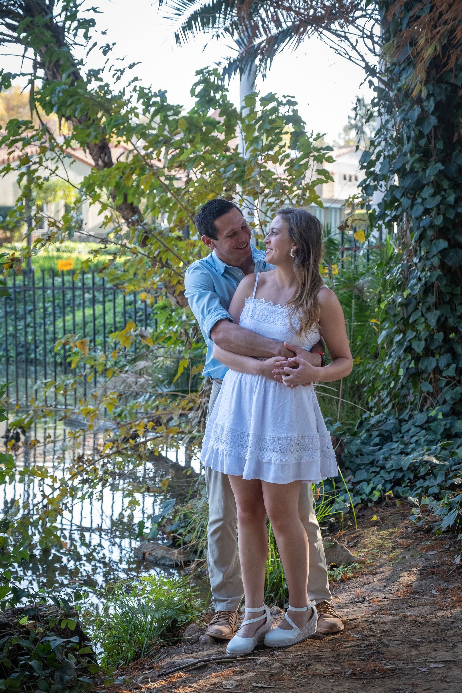 Man embracing woman in a white sundress near a pond in a lush, ivy-filled garden setting.