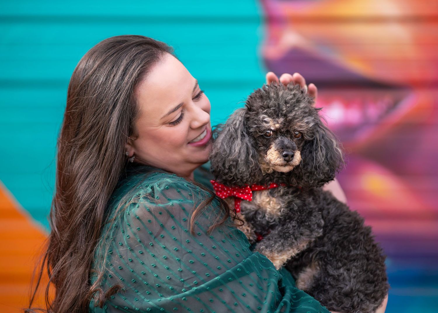 A warm embrace between a person wearing teal and a curly-haired dog against a colorful background.
