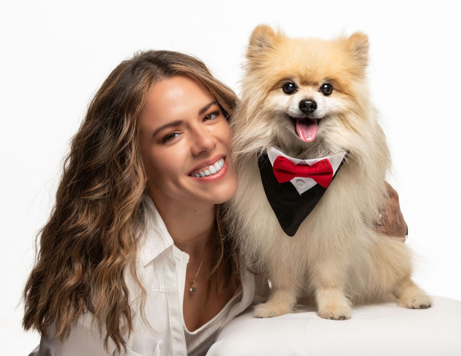 Smiling woman with long brown hair poses with a fluffy Pomeranian dog wearing a tuxedo and red bow tie.