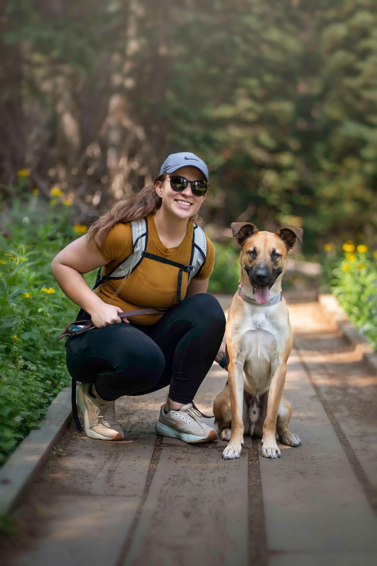 Woman in hiking gear crouches beside a tan dog on a wooden trail path surrounded by green foliage.