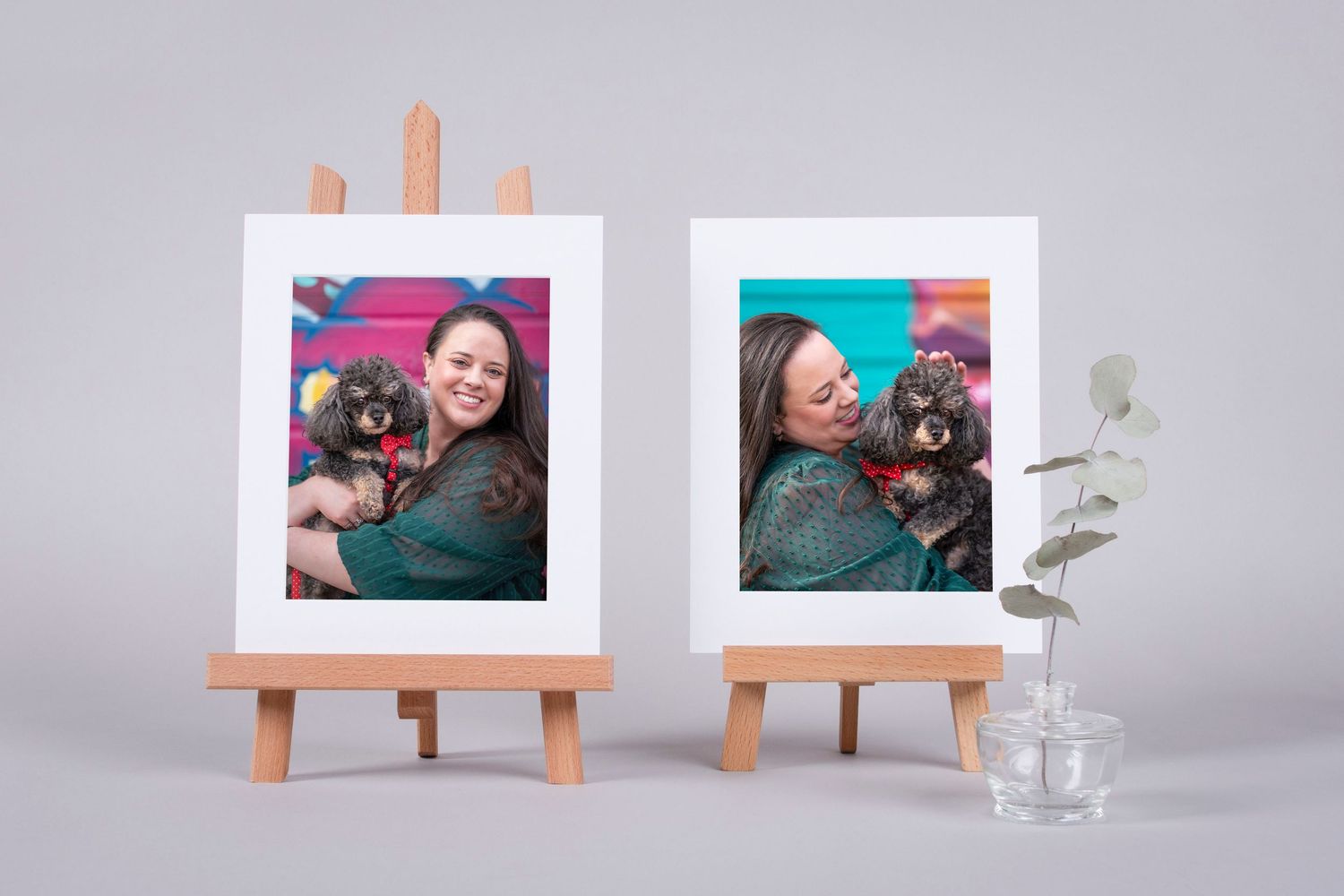 Two framed photos of a smiling woman holding a small dog displayed on wooden easels beside a glass vase with eucalyptus.