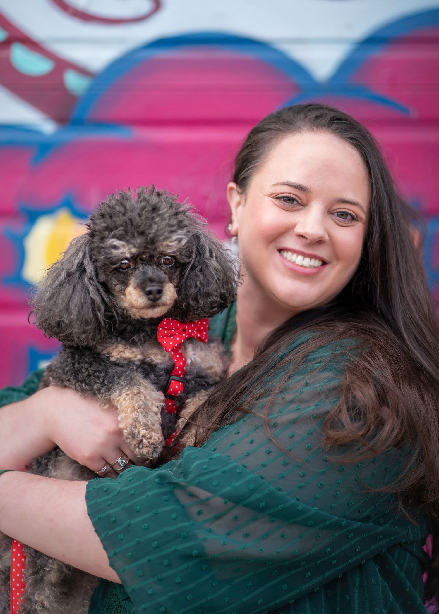 A small curly-haired poodle mix with a red bowtie poses with someone against a colorful graffiti background.